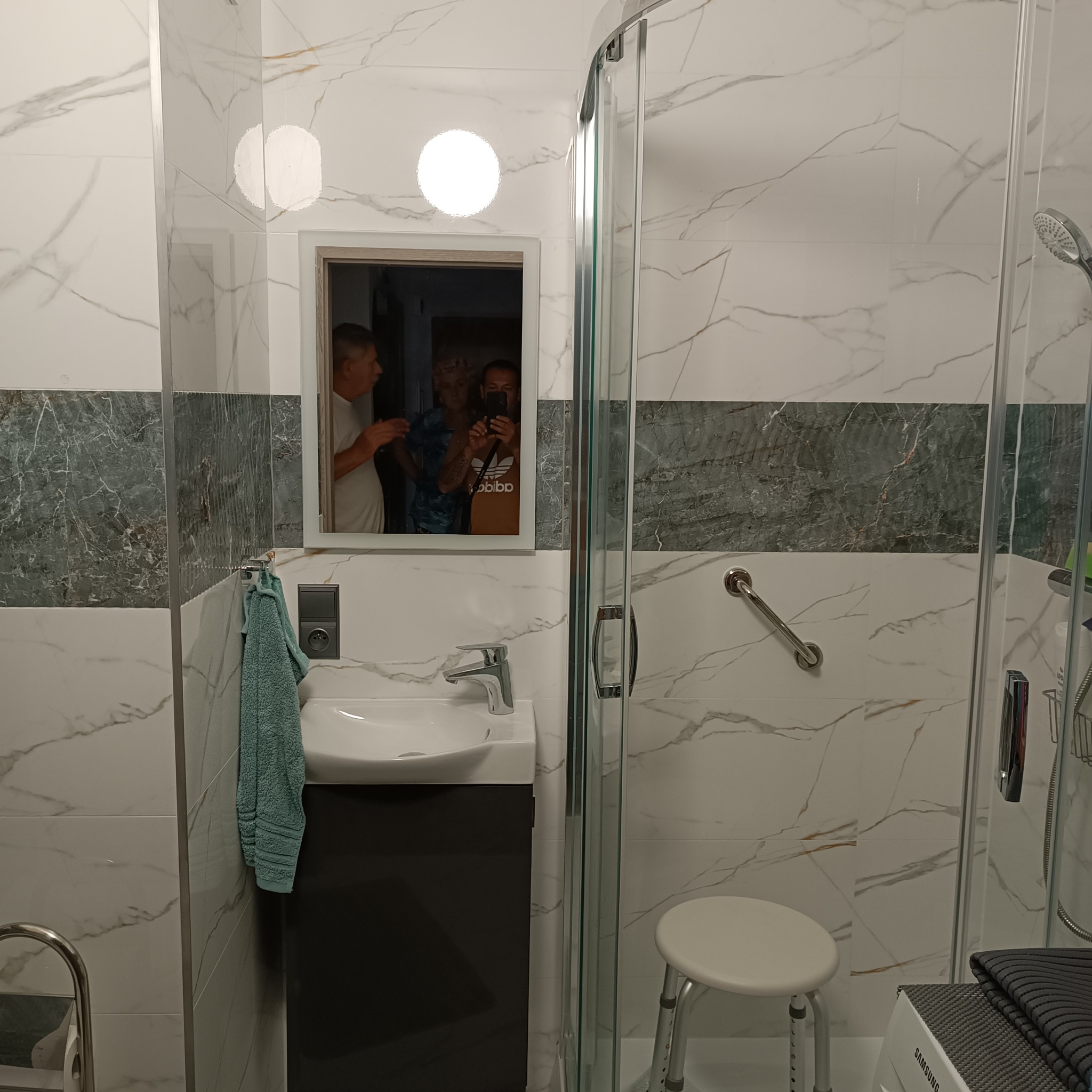 Wykończona łazienka z kabiną prysznicową, umywalką z szafką w kolorze antracytowym, biało-szarymi płytkami imitującymi marmur i akcentem z płytek w odcieniach zieleni, z widocznym odbiciem trzech...