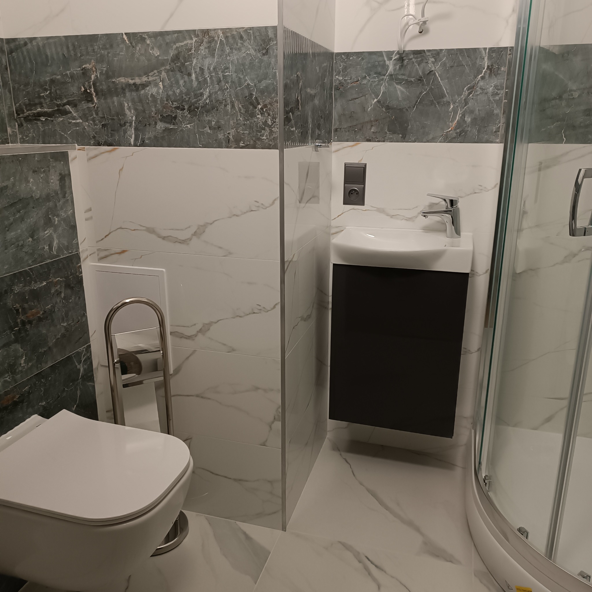 Nowoczesna łazienka z marmurowymi płytkami w odcieniach bieli i szarości, wisząca toaleta z chromowanym uchwytem na papier, szafka pod umywalką w kolorze grafitowym oraz kabina prysznicowa...