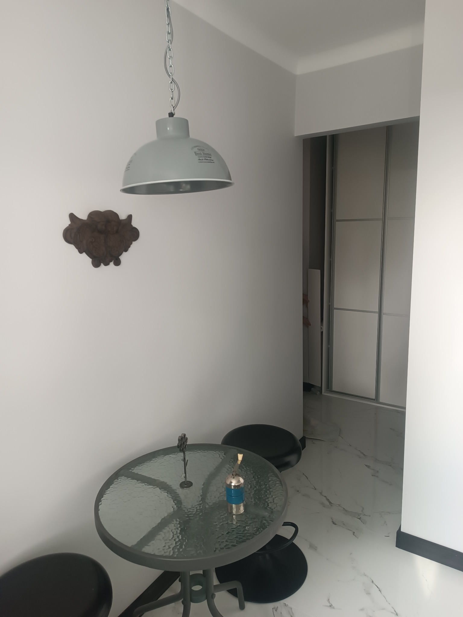 Szklany stół z metalową ramą i dwoma czarnymi stołkami w rogu pokoju z marmurową podłogą, wisząca szara lampa nad stołem, ozdobny element na ścianie i biała szafa z przesuwnymi drzwiami w tle.