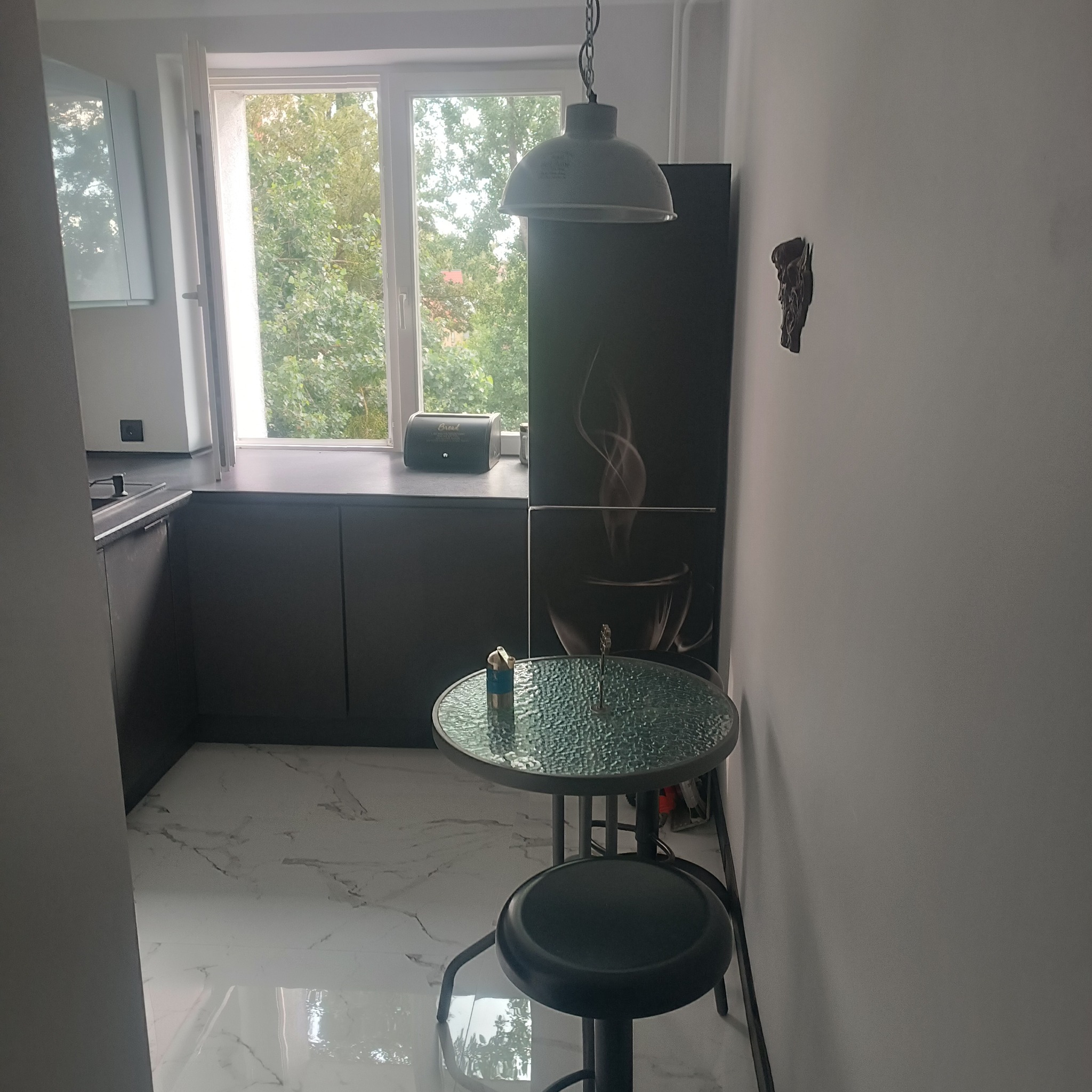 Nowoczesna kuchnia z czarnymi szafkami, marmurową podłogą, okrągłym szklanym stolikiem i wiszącą lampą w stylu industrialnym; na ścianie dekoracja przypominająca rzeźbę.