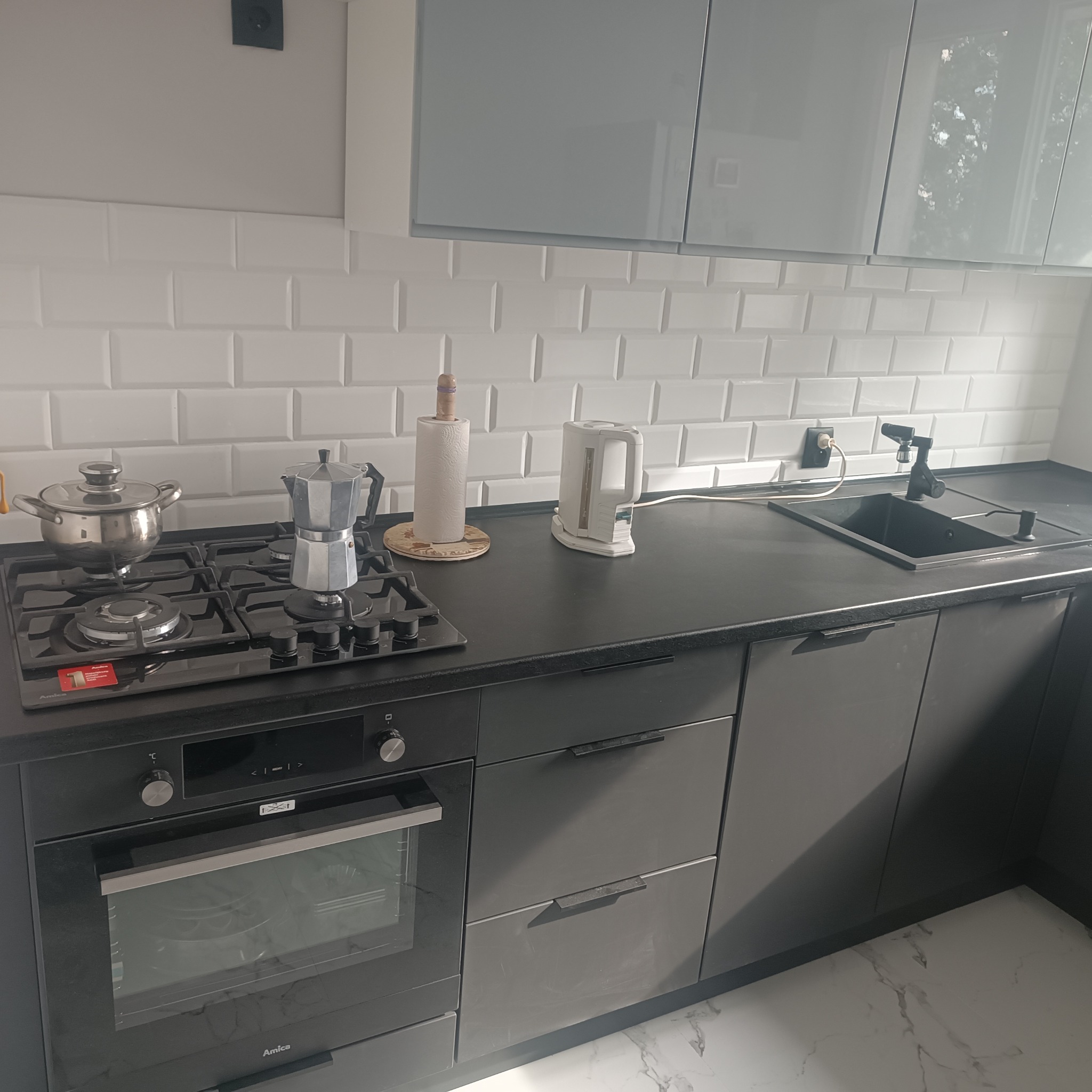 Nowoczesna, szara kuchnia z białymi płytkami typu metro, czarnym blatem i sprzętem AGD: piekarnik Amica, płyta gazowa, czarny zlew i bateria. Na blacie czajnik i kawiarka.