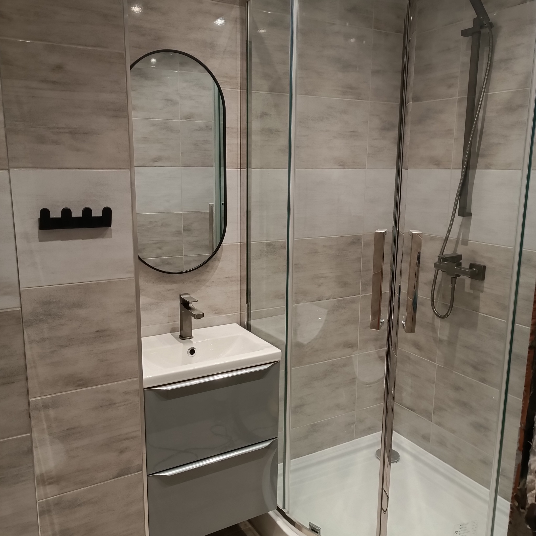 Nowoczesna łazienka z kabiną prysznicową ze szklanymi drzwiami, umywalką z szarą szafką, owalnym lustrem i płytkami imitującymi kamień na ścianach.