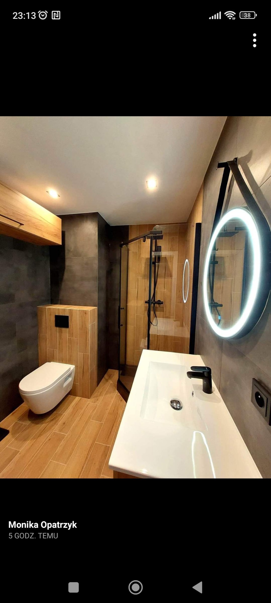 Nowoczesna łazienka z drewnopodobnymi płytkami, podwieszaną toaletą, kabiną prysznicową i okrągłym lustrem z podświetleniem LED.