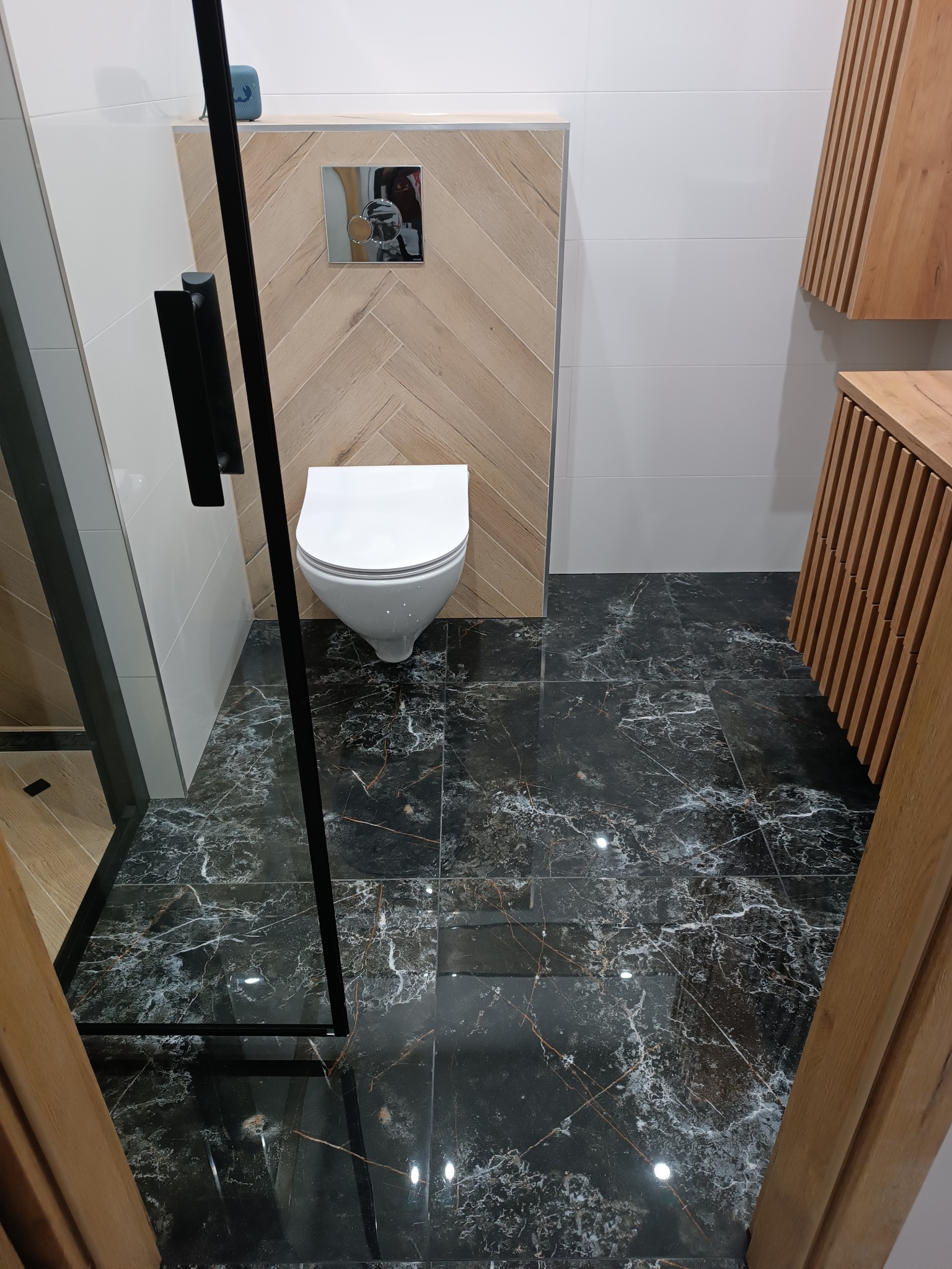 Nowoczesna łazienka z podwieszaną toaletą, czarną marmurową podłogą, drewnianą ścianką z mozaiką w jodełkę i szafką z lamelami.