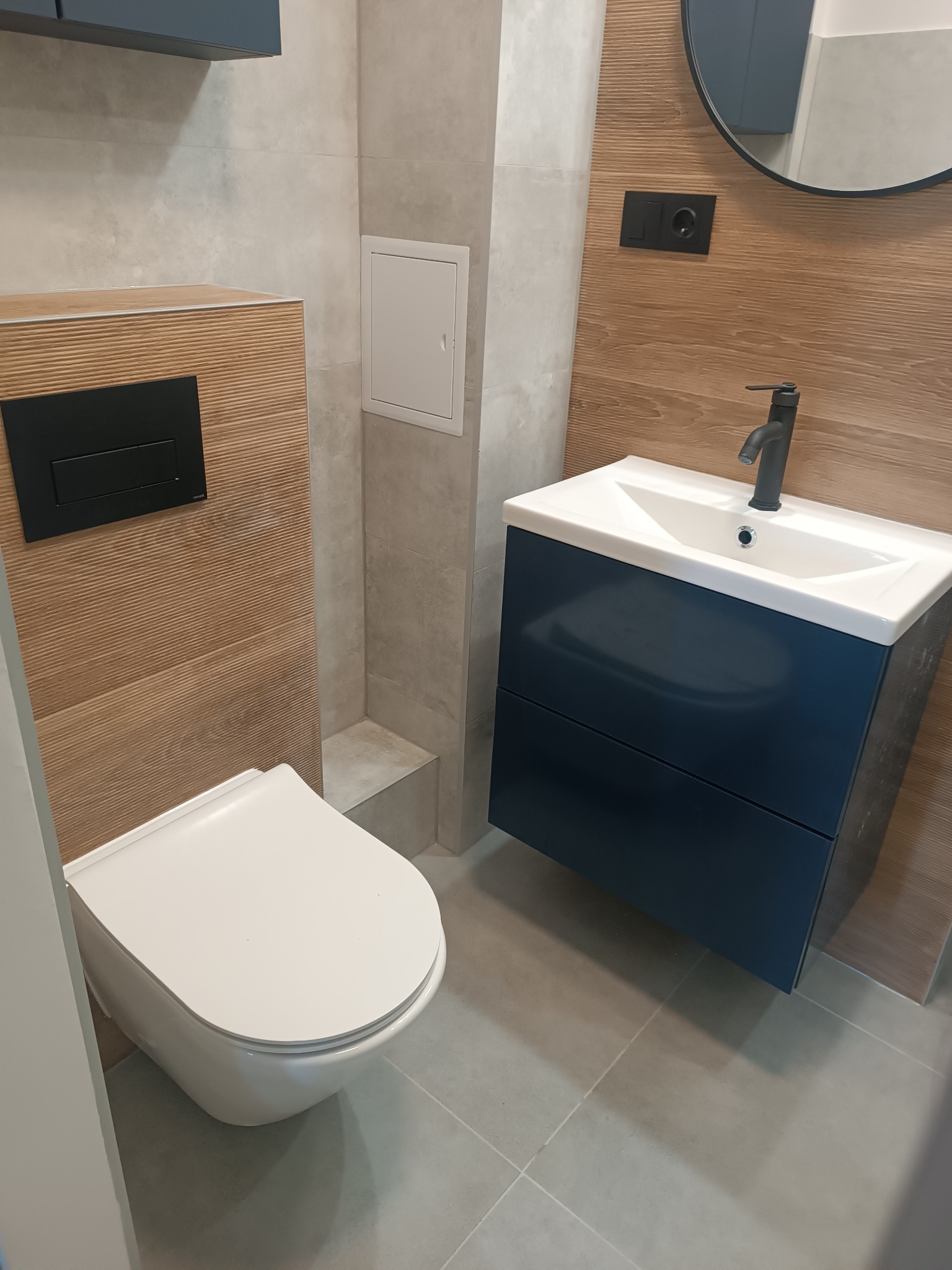 Nowoczesna łazienka z podwieszaną toaletą, umywalką w szafce w kolorze granatowym i drewnopodobnymi płytkami na ścianie, uzupełniona o okrągłe lustro i czarną armaturę.