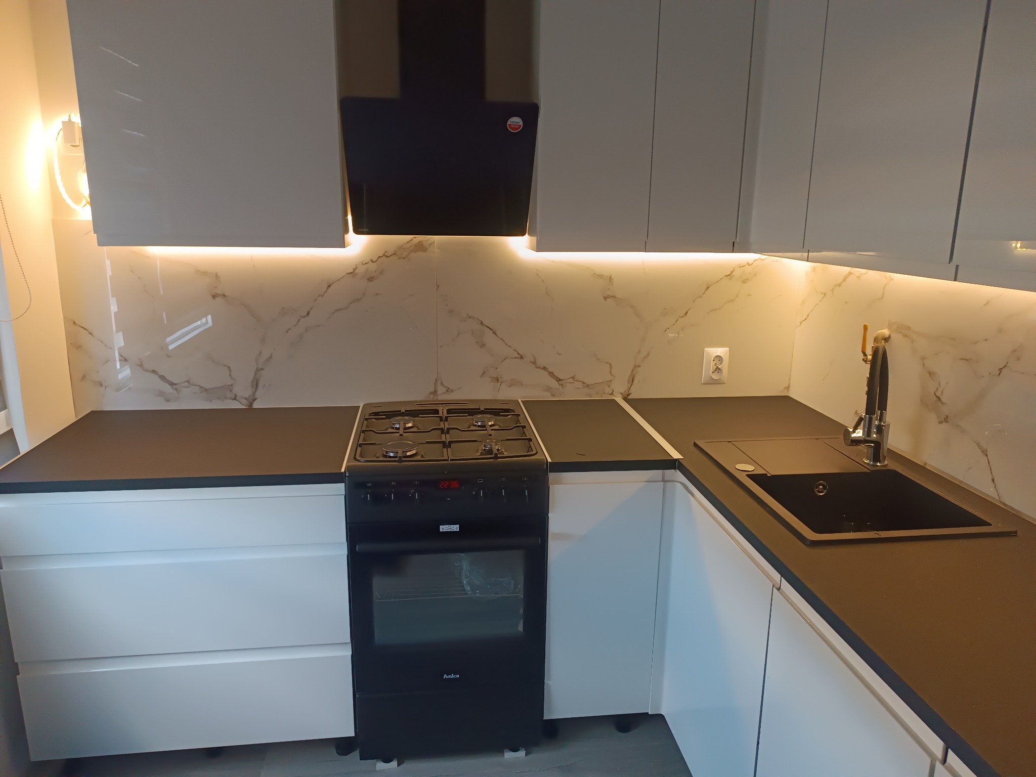 Nowoczesna kuchnia z białymi szafkami, czarnym blatem i płytą gazową Amica, oświetleniem LED pod szafkami wiszącymi i marmurowym wzorem na ścianie.