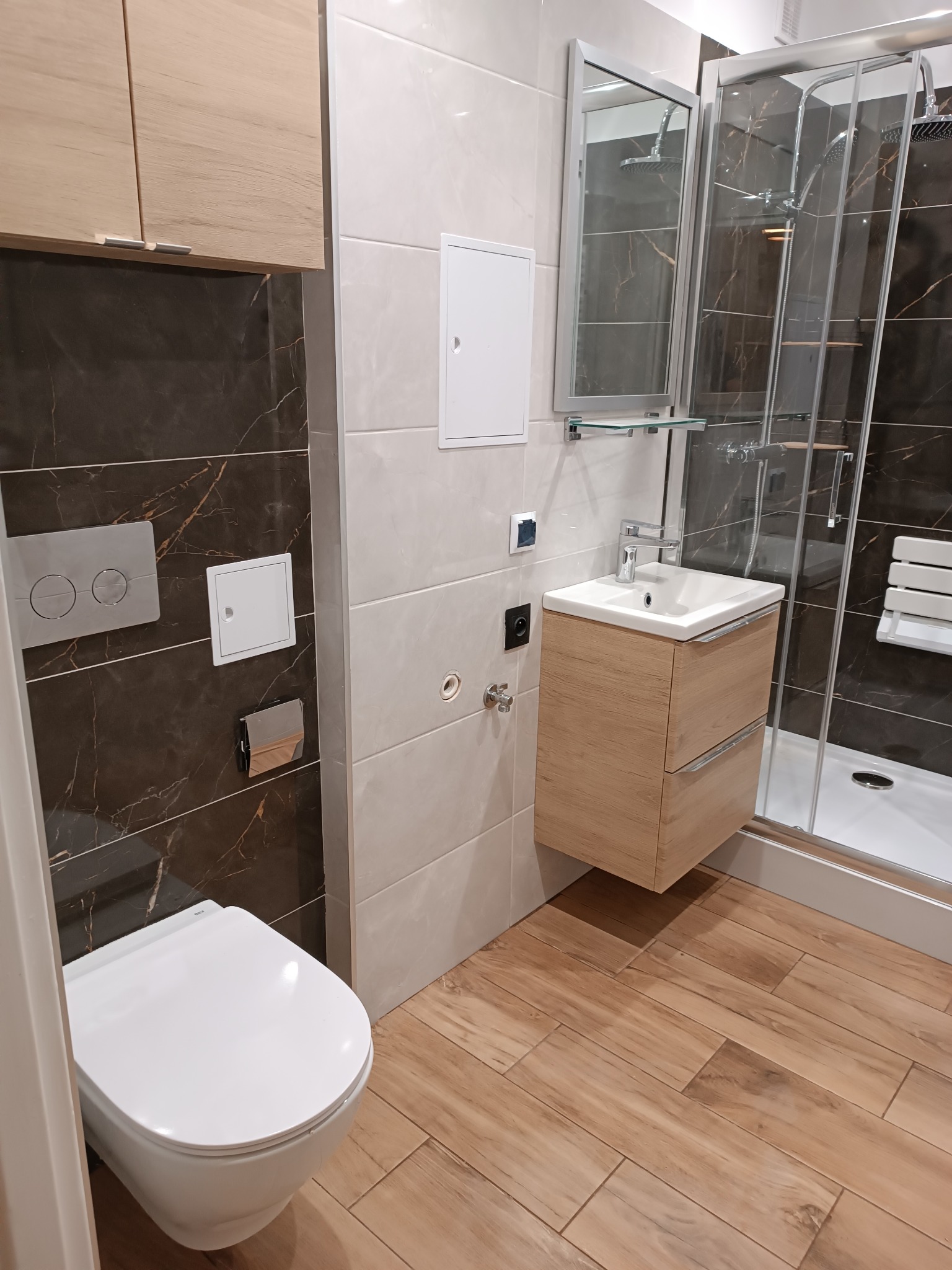 Nowoczesna łazienka z drewnopodobną podłogą, brązowymi i beżowymi płytkami, podwieszaną toaletą, szafką z umywalką z drewnianym frontem oraz kabiną prysznicową z siedziskiem.