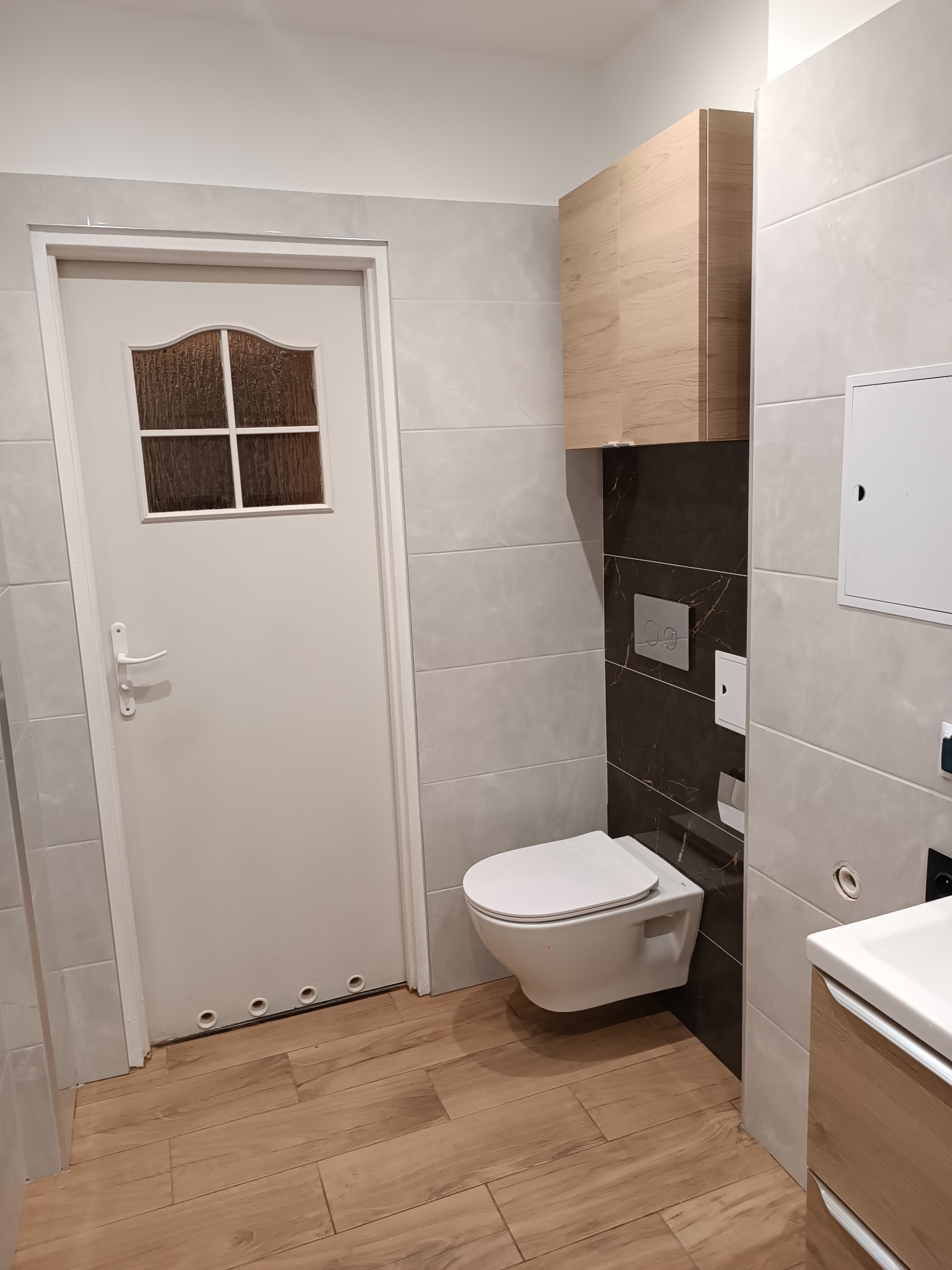 Nowoczesna łazienka w jasnych szarościach z akcentami drewna: wisząca toaleta, szafka nad nią i drewnopodobna podłoga, białe drzwi z szybą w kratkę po lewej stronie.