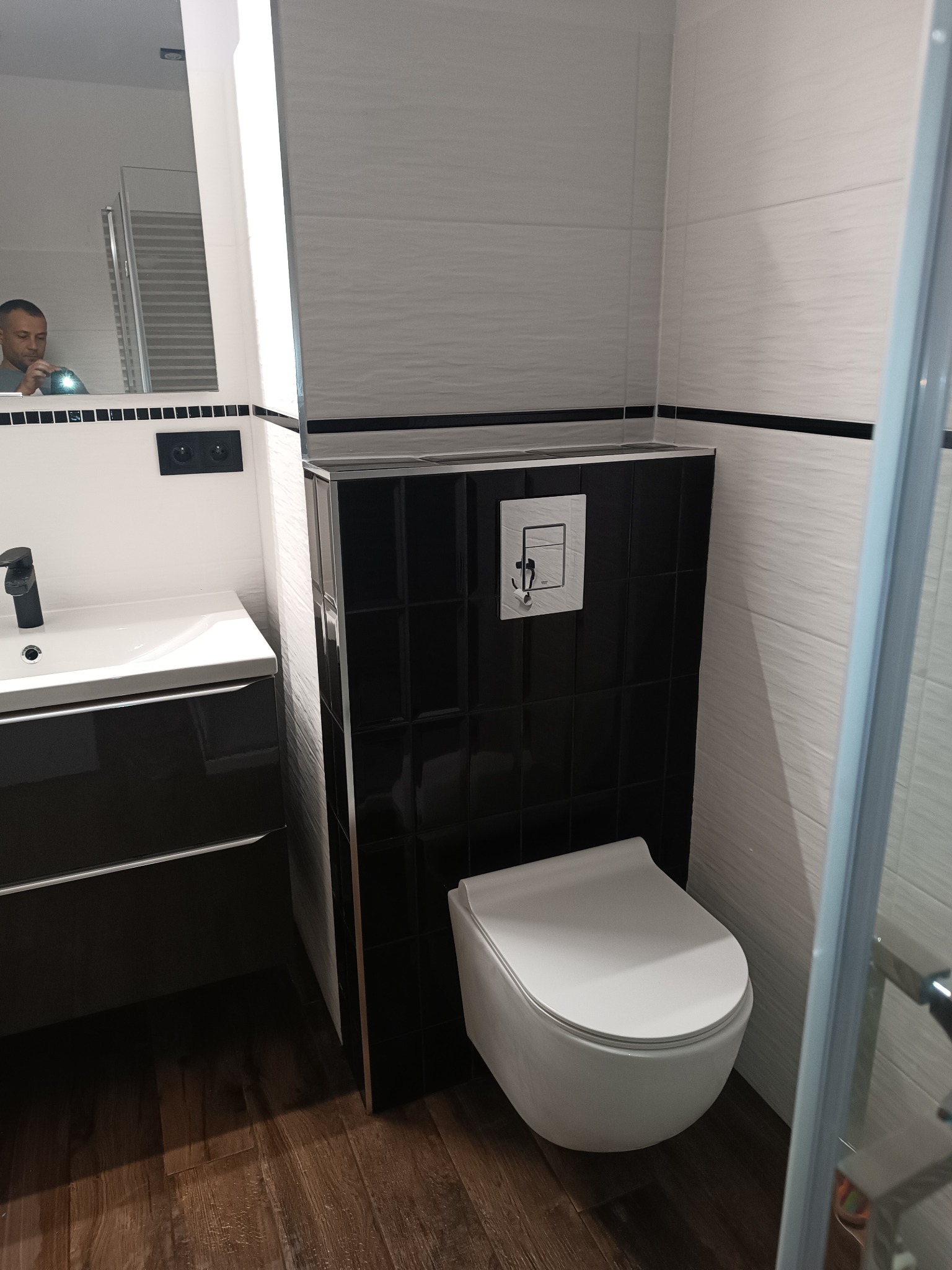 Nowoczesna łazienka z podwieszaną miską WC, czarną zabudową stelaża z płytek i umywalką z szafką w ciemnym kolorze, widok z perspektywy kabiny prysznicowej, odbicie osoby w lustrze.