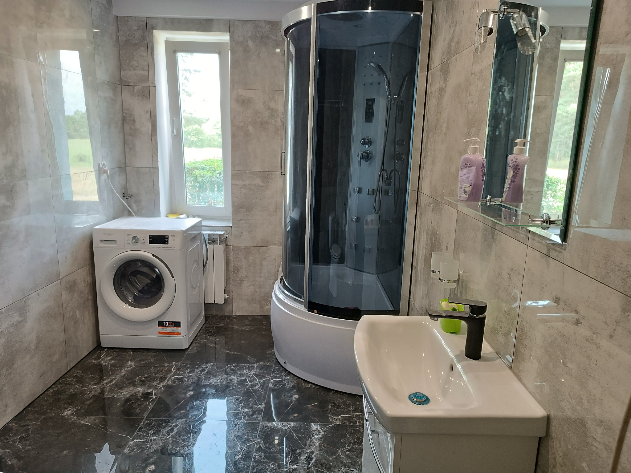 Nowoczesna łazienka z kabiną prysznicową z hydromasażem, umywalką na postumencie, pralką i szarymi płytkami imitującymi beton, wykończona w chłodnej tonacji.