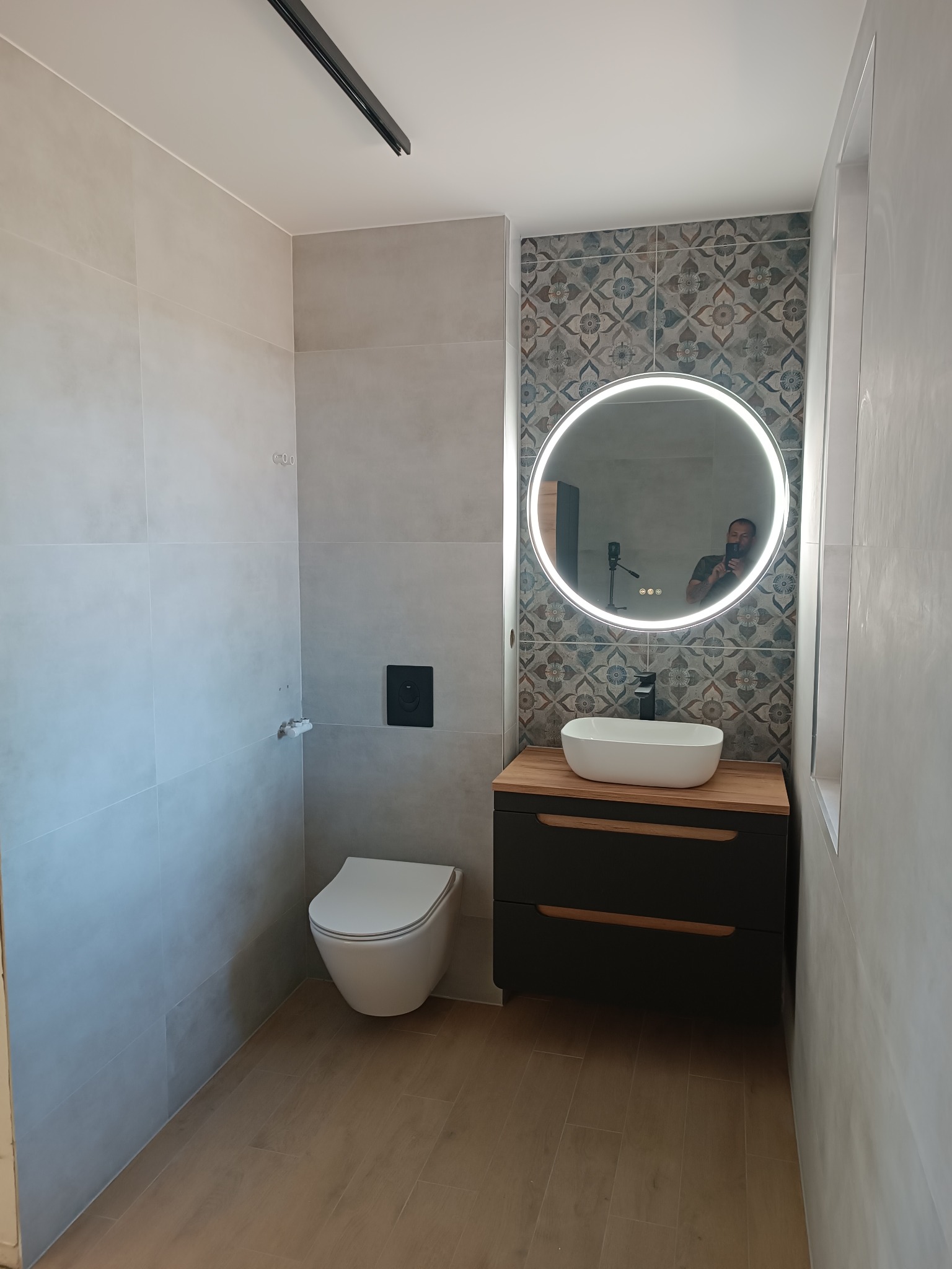 Nowoczesna łazienka z szarymi płytkami, okrągłym lustrem z podświetleniem LED, białą umywalką nablatową, czarną szafką pod umywalką z drewnianym blatem i podwieszaną toaletą.
