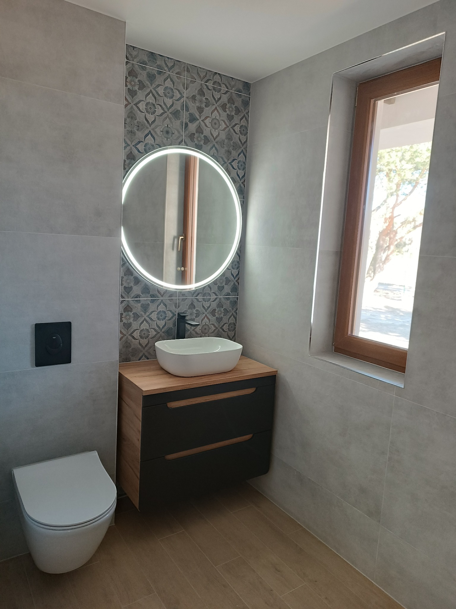 Nowoczesna łazienka z szarymi płytkami, okrągłym lustrem z podświetleniem LED nad białą umywalką nablatową, drewnianą szafką z ciemnymi frontami i wiszącą toaletą.