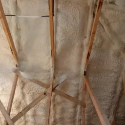 Poddasze z drewnianą konstrukcją krokwiową wypełnione beżową pianką izolacyjną o nieregularnej strukturze; widoczne ślady montażu instalacji.