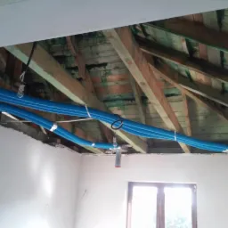 Niebieskie, karbowane rury wentylacyjne przymocowane do drewnianej konstrukcji stropu w surowym stanie, widoczne elementy montażowe i okno w tle.