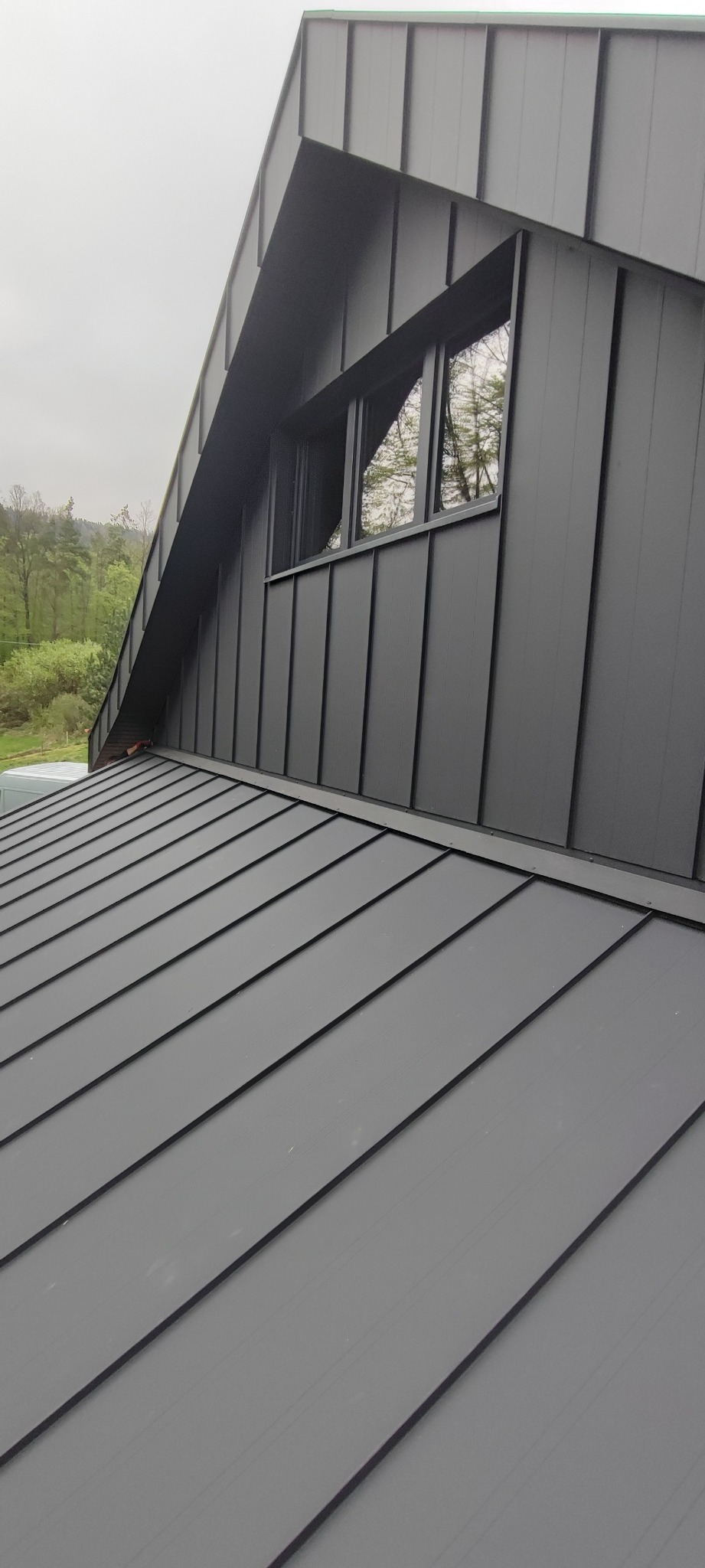 Nowoczesny, szary dach z blachy na budynku z oknem, widoczne panele blaszane ułożone pod kątem, w tle zieleń drzew.