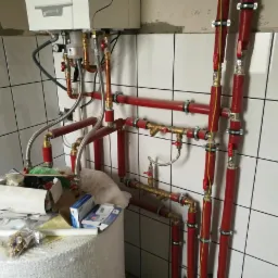 Nowa instalacja grzewcza z piecem De Dietrich, rury w czerwonej izolacji, widoczne zawory i przyłącza, na pierwszym planie biały zbiornik z narzędziami i materiałami montażowymi.