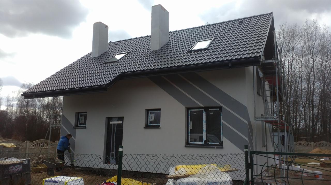Dwukondygnacyjny dom z ciemną dachówką i szarymi pasami na elewacji w trakcie wykończenia; widoczne rusztowania, okna z folią ochronną i pracownik przy wyrównywaniu terenu.