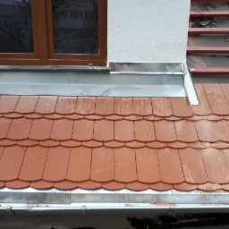 Nowe, pomarańczowo-czerwone dachówki ceramiczne ułożone na połaci dachu, z widocznymi obróbkami blacharskimi przy oknie i rynną. Część dachu jeszcze bez pokrycia, widoczna konstrukcja drewniana.