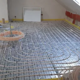 Rozłożone rury ogrzewania podłogowego na siatce w trakcie montażu w pomieszczeniu na poddaszu, widoczne narzędzia i zwój rur.