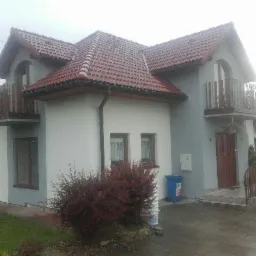 Dwukondygnacyjny dom z czerwonym dachem, balkonami z drewnianą balustradą i elewacją w odcieniach szarości i bieli, widoczny z zewnątrz, z krzewem przed wejściem i niebieskim pojemnikiem na śmieci...