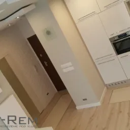 Nowoczesna kuchnia z białymi szafkami, wbudowanym piekarnikiem, drewnianą podłogą i dekoracyjnym sufitem z oświetleniem punktowym, widoczny zegar na ścianie i logo firmy w rogu zdjęcia.