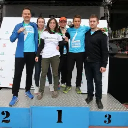 Grupa osób stojąca na podium z numerami startowymi i medalami, z logo Oferteo.pl na koszulce jednej z osób, na tle sceny z trofeami i kartonem pralki.