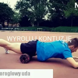 Mężczyzna w niebieskiej koszulce roluje mięsień czworogłowy uda za pomocą rollera na zewnątrz, na tle drzew i bieżni.