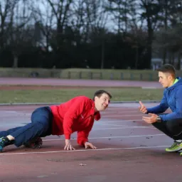 Mężczyzna w czerwonej bluzie ćwiczy z rollerem na bieżni, drugi mężczyzna w niebieskiej bluzie kuca obok i udziela instrukcji.