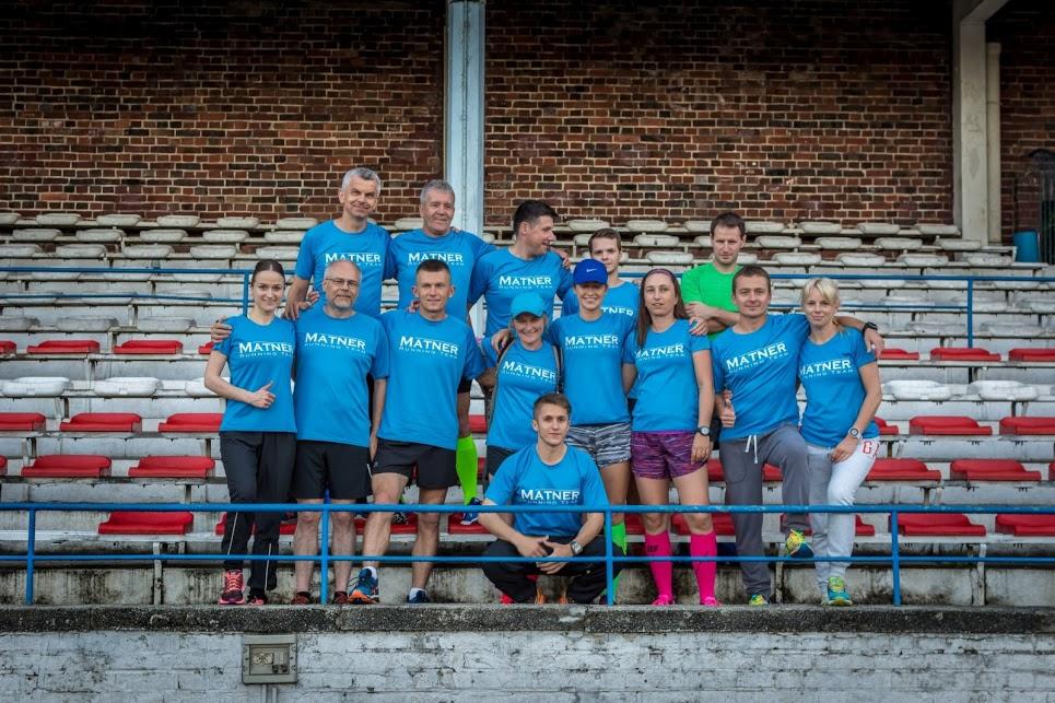 Grupa kilkunastu osób w niebieskich koszulkach z napisem 'MATNER RUNNING TEAM' pozuje na tle trybun stadionu z ceglaną ścianą w tle.