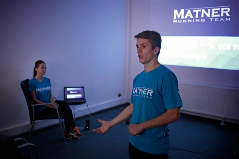 Młody mężczyzna w niebieskiej koszulce z logo 'MATNER RUNNING TEAM' prezentuje coś na tle wyświetlanego ekranu z informacjami kontaktowymi, w tle siedzi kobieta również w koszulce z logo.