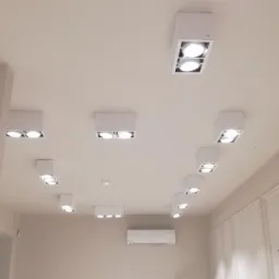 Białe, kwadratowe oprawy oświetleniowe z dwoma żarówkami LED, rozmieszczone symetrycznie na suficie w jasnym pomieszczeniu z klimatyzacją.