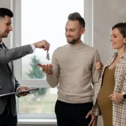 Agent nieruchomości wręcza klucze uśmiechniętej parze, kobieta w ciąży, w tle okno i logo firmy Kurapiewicz Nieruchomości.