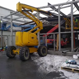 Montaż konstrukcji stalowej budynku, pracownicy na podnośnikach koszowych w trakcie łączenia elementów, widoczne spawane połączenia