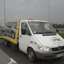 Biała laweta Mercedes z uszkodzonym srebrnym samochodem osobowym na żółtej platformie, widoczny numer telefonu i logo firmy Smart Truck na masce pojazdu, szara nawierzchnia drogi i pochmurne niebo...