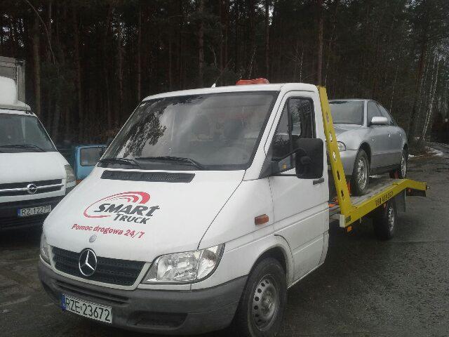 Biała laweta Mercedes-Benz z logo 'SMART TRUCK Pomoc drogowa 24/7' przewozi srebrny samochód osobowy na żółtej platformie, na tle ciemnego lasu.