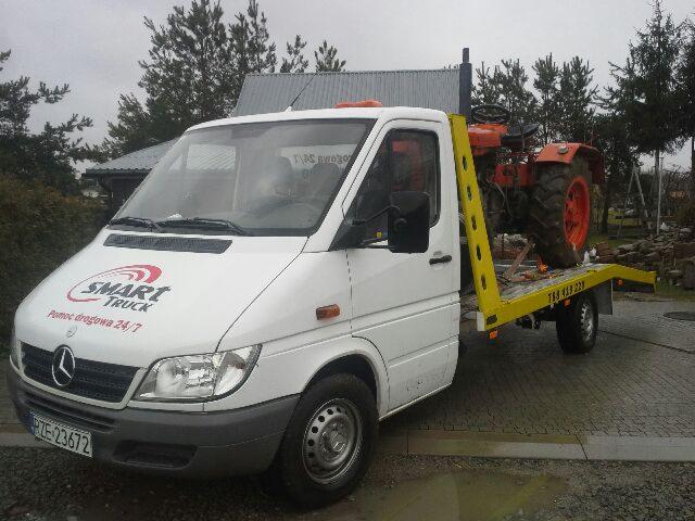 Biała laweta z żółtym najazdem przewozi czerwony traktor rolniczy na tle szarej kostki brukowej i zielonych drzew