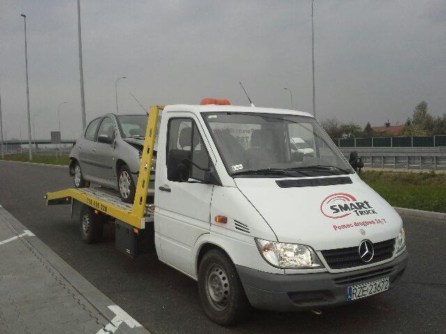Biała laweta Mercedes z uszkodzonym srebrnym samochodem osobowym na żółtej platformie, widoczny numer telefonu i logo firmy Smart Truck na masce pojazdu, szara nawierzchnia drogi i pochmurne niebo...