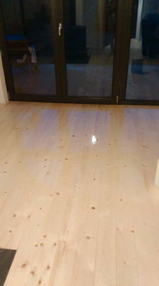 Jasne drewniane deski podłogowe z widocznymi sękami, świeżo polakierowane, z refleksami światła, na tle ciemnych drzwi tarasowych.