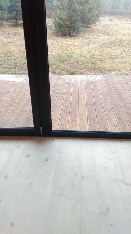 Jasne deski podłogowe wewnątrz, przechodzące w drewniany taras na zewnątrz, widok przez czarne, przesuwne drzwi tarasowe, na zewnątrz trawa i drzewa.