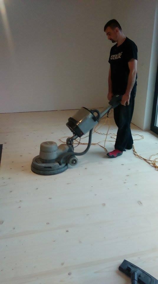 Mężczyzna w czarnej koszulce obsługuje maszynę do renowacji jasnej drewnianej podłogi w pomieszczeniu z białymi ścianami, widoczny kabel zasilający i fragment okna.