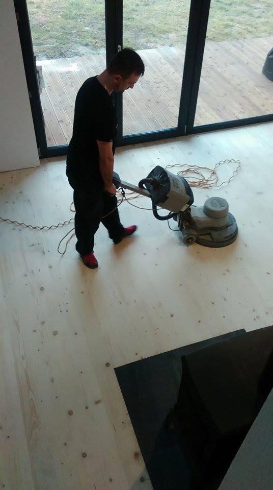 Mężczyzna w czarnym ubraniu i czerwonych butach cyklinuje drewnianą podłogę za pomocą profesjonalnej maszyny do cyklinowania w przestronnym wnętrzu z dużymi oknami.