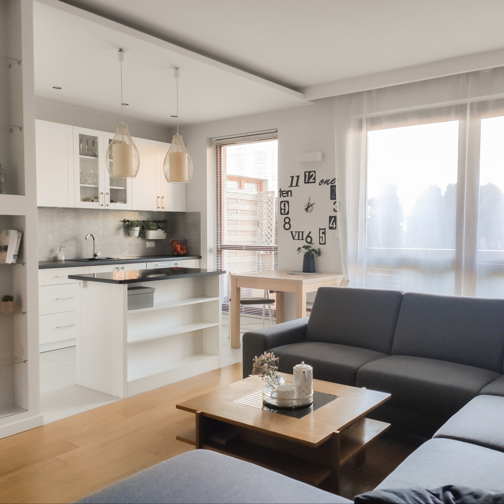 Jasne wnętrze mieszkania z aneksem kuchennym, szarą sofą narożną i drewnianym stolikiem kawowym z dekoracjami.