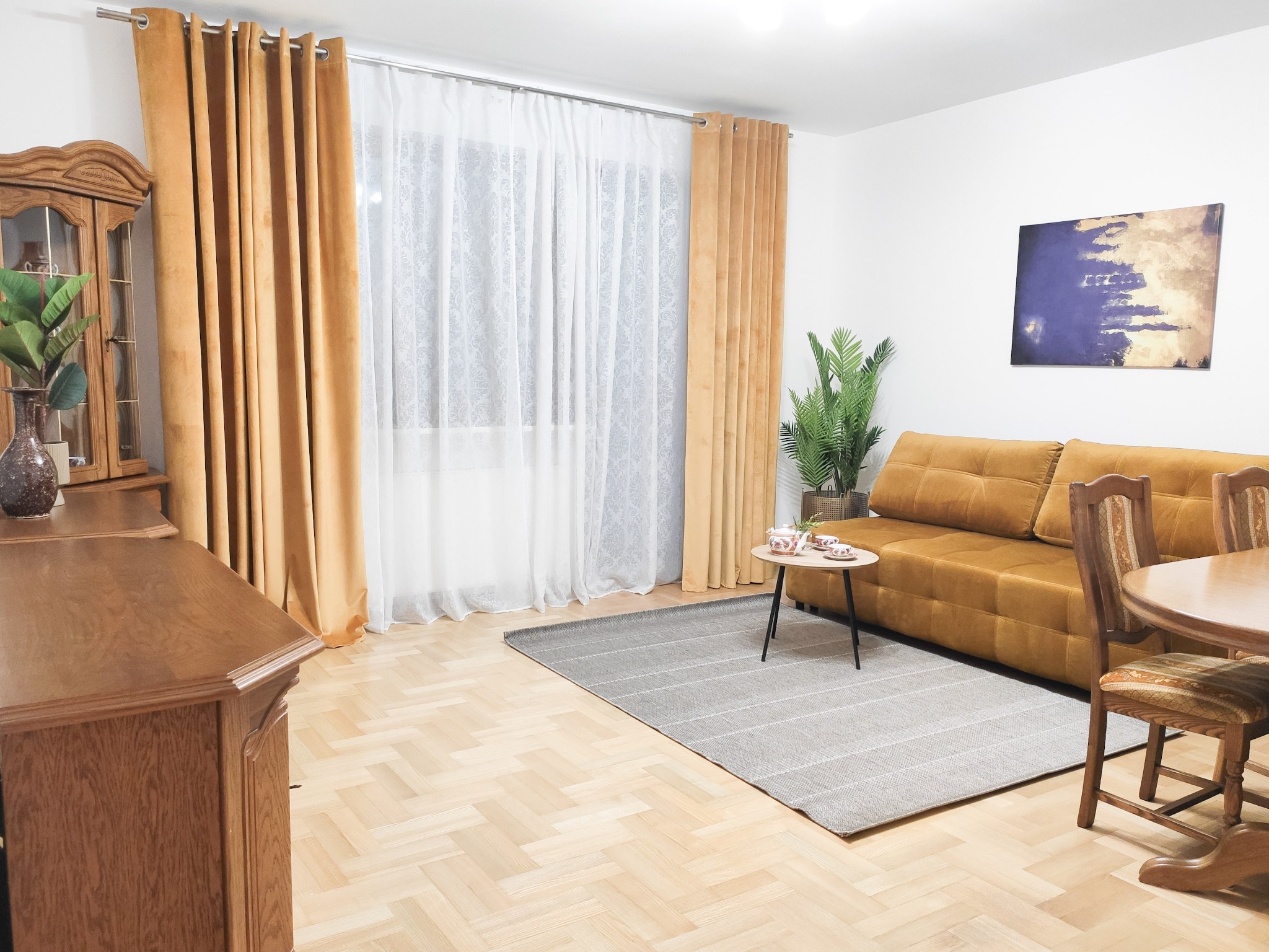 Jasne wnętrze salonu z parkietem w jodełkę, musztardową sofą, zasłonami w tym samym odcieniu i drewnianymi meblami w stylu retro.