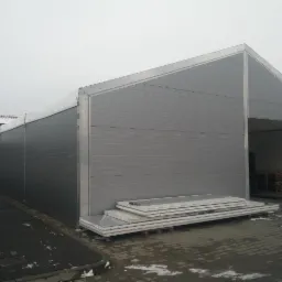 Szara hala magazynowa z konstrukcji stalowej z widocznymi panelami warstwowymi na palecie przed budynkiem, pochmurny dzień.