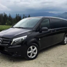 Czarny van Mercedes-Benz Vito na szutrowej drodze z widokiem na zalesione wzgórza i góry w tle, pochmurne niebo.