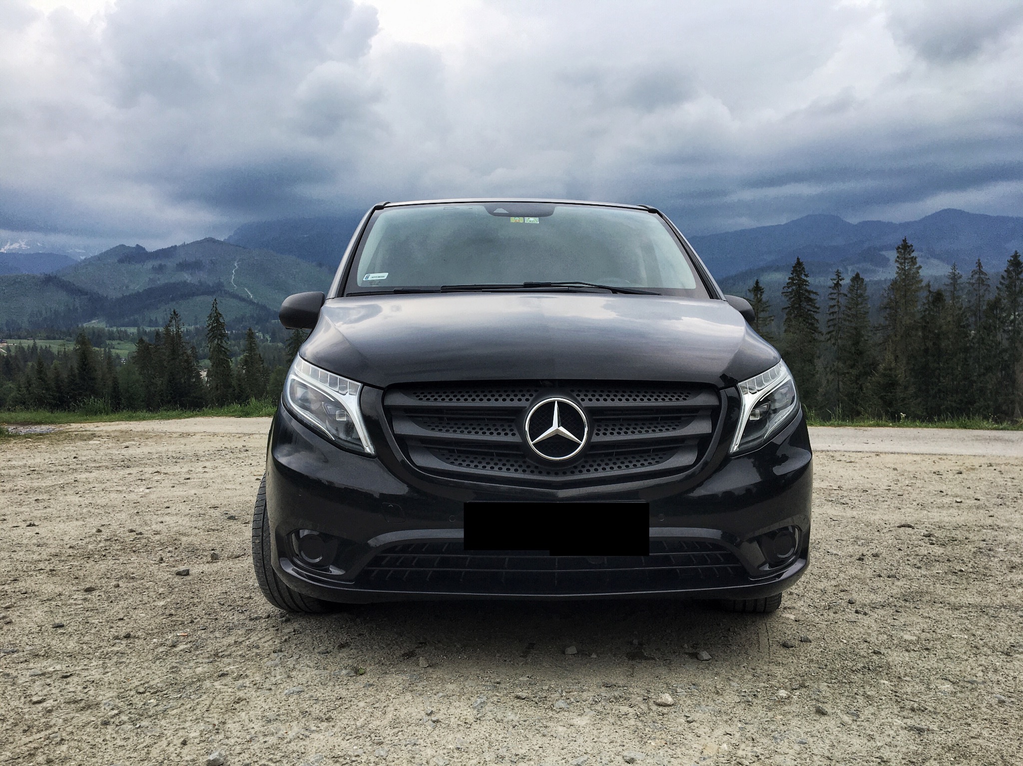 Czarny Mercedes Vito na szutrowej drodze z widokiem na zalesione góry i pochmurne niebo w tle.