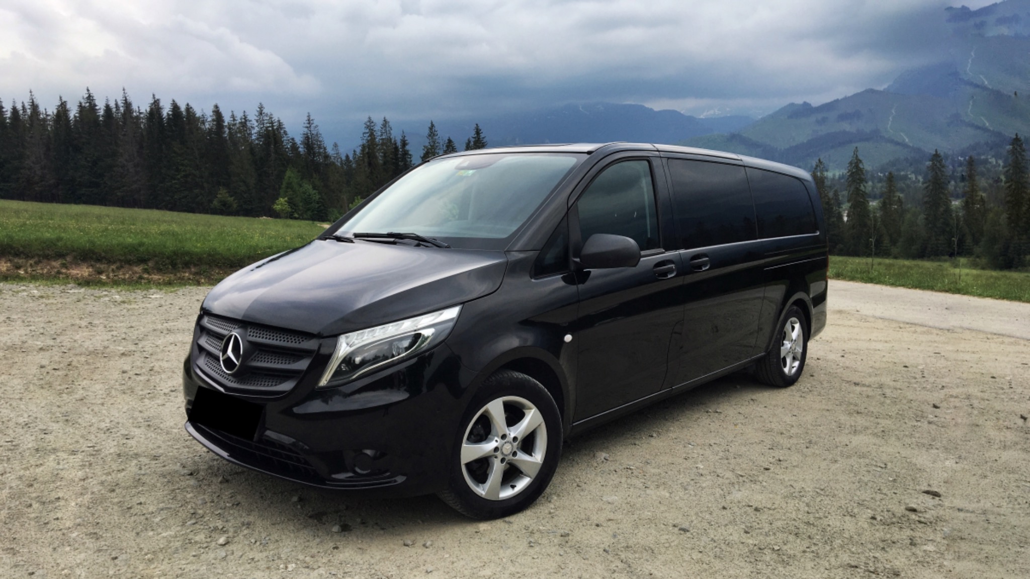 Czarny van Mercedes-Benz Vito na szutrowej drodze z widokiem na zalesione wzgórza i góry w tle, pochmurne niebo.
