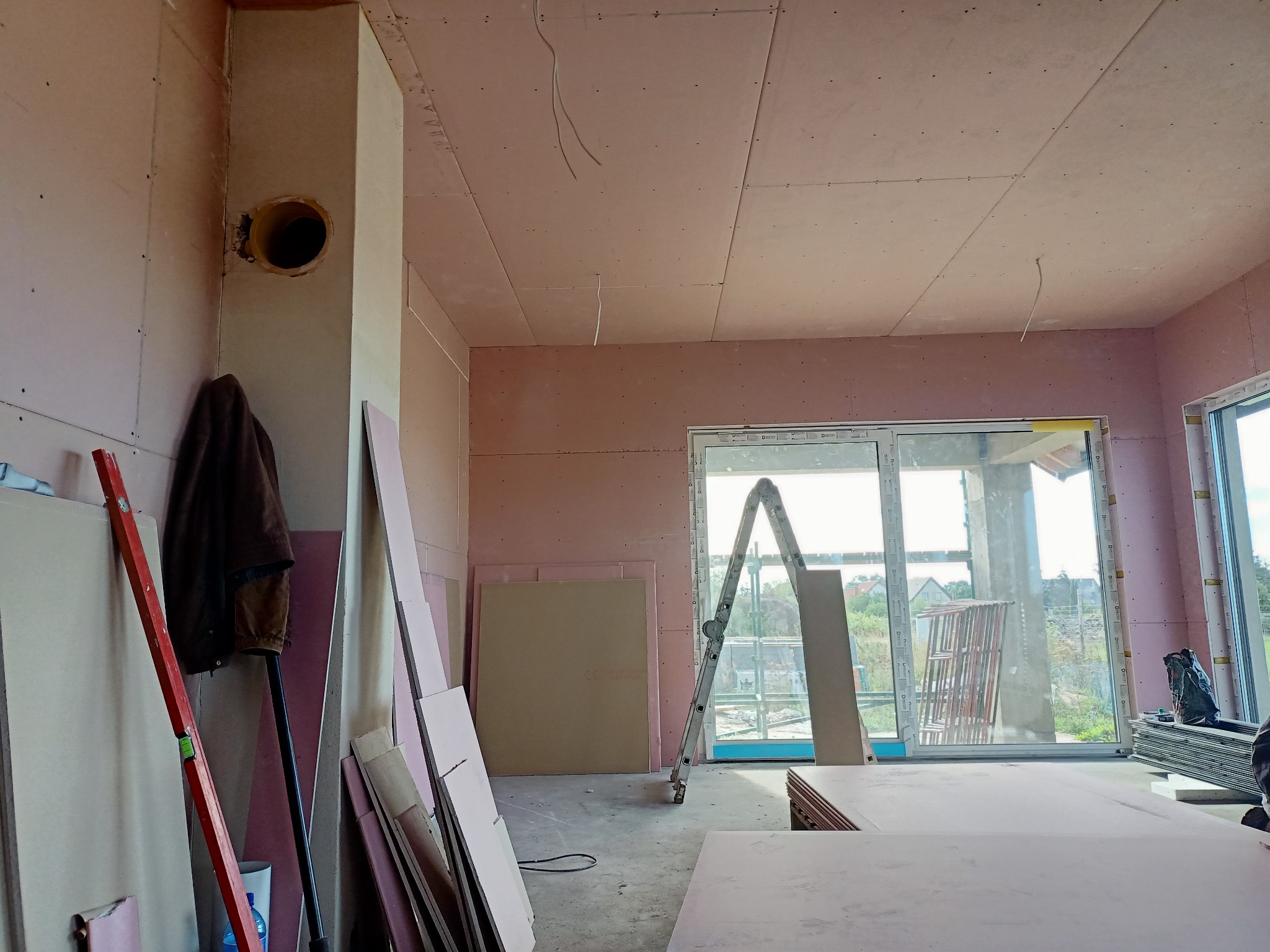Wnętrze w trakcie montażu płyt kartonowo-gipsowych na ścianach i suficie, widoczne łączenia płyt, drabina oparta o okno balkonowe oraz stos płyt. W rogu pokoju widoczna pionowa rura wentylacyjna...