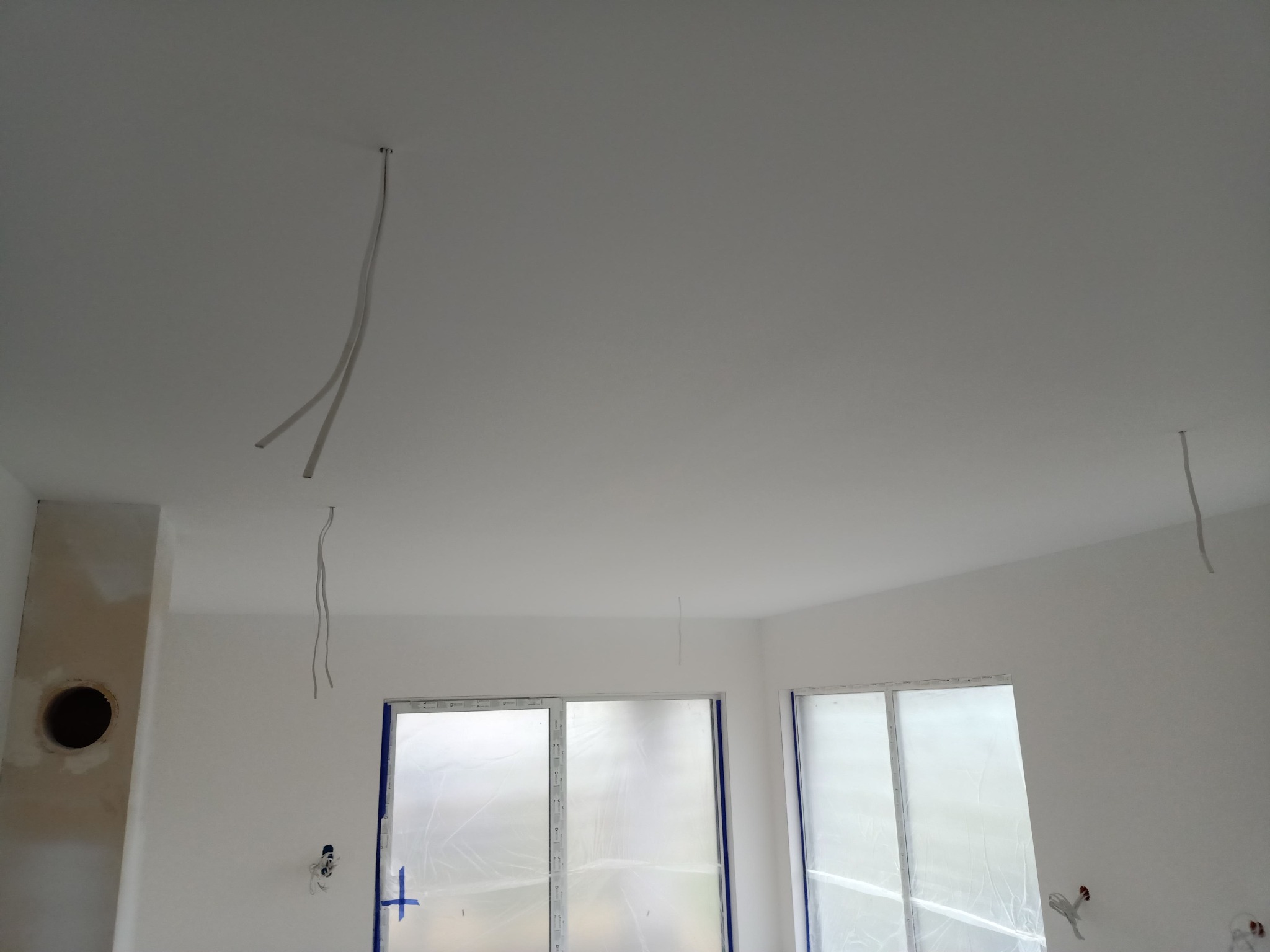 Wnętrze w trakcie remontu: biały sufit z wiszącymi kablami elektrycznymi, ściany przygotowane pod malowanie, okna zabezpieczone folią ochronną.