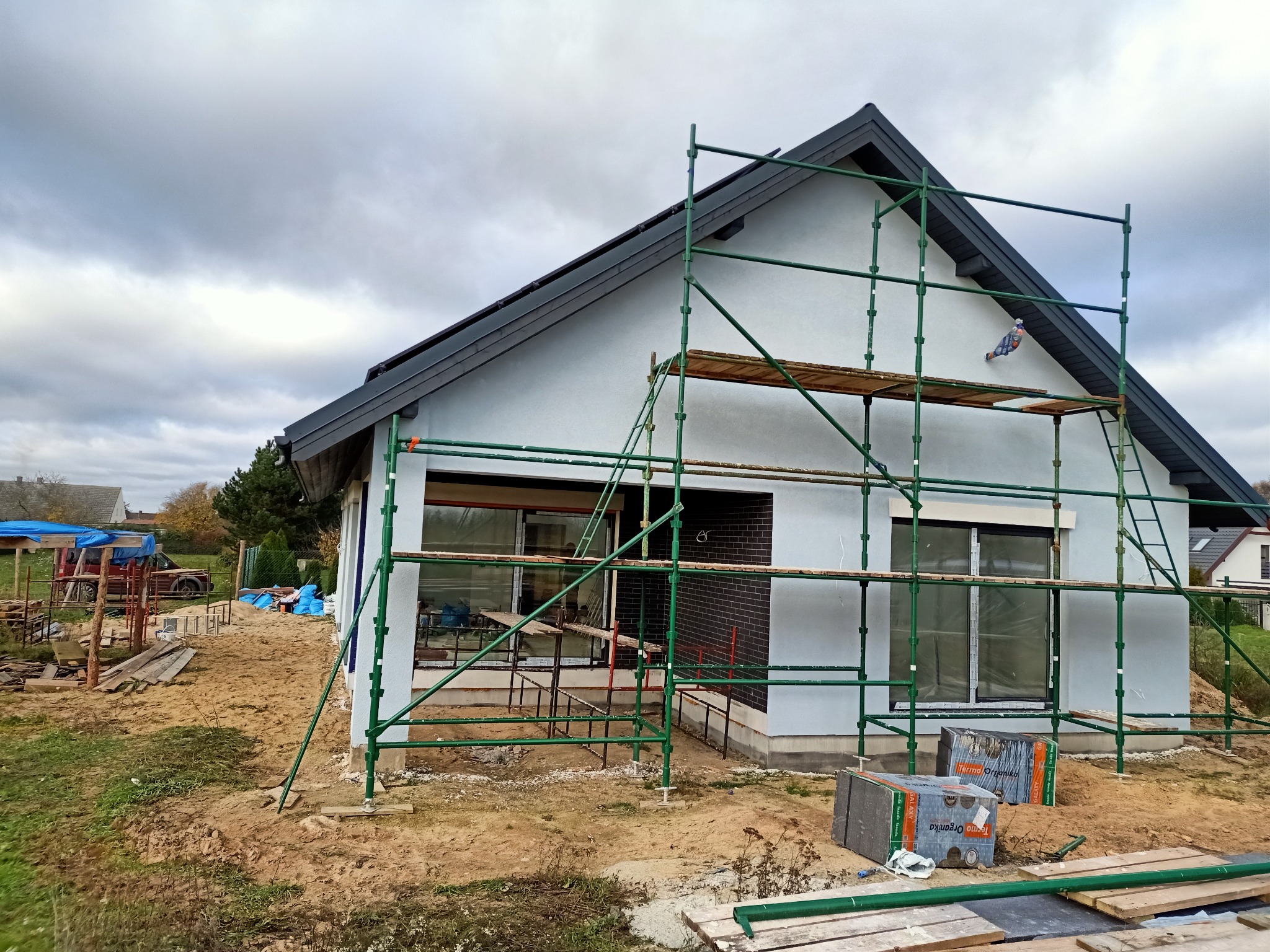 Dom jednorodzinny w trakcie prac elewacyjnych, widoczne rusztowania, częściowo otynkowana ściana i materiały izolacyjne na ziemi.